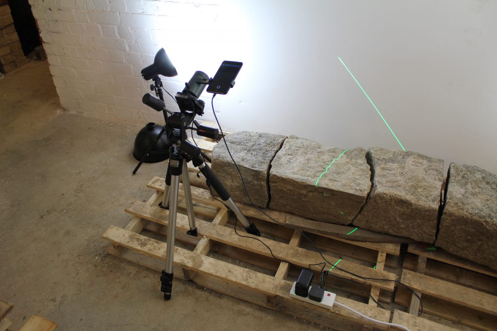 A large stone broken into three pieces lays upon a wooden pallet. On a tripod is a 3D laser scanner, which is capturing the surface of the stone with a green laser stripe which is visible sweeping across the surface of the stone fragments